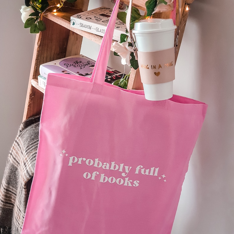 May include: Pink tote bag with the text "probably full of books" in white lettering. The bag is hanging on a wooden shelf with books and a coffee mug with a brown sleeve that says "mug in a mug" in gold lettering.