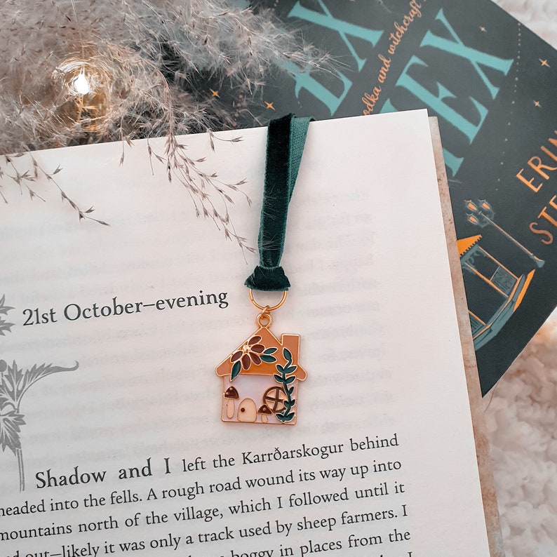 May include: A gold-toned bookmark with a green velvet ribbon. The bookmark features a small, detailed house with a brown roof, white walls, and a green vine. The house has a small window and a door with a brown frame. There are mushrooms and flowers around the house.