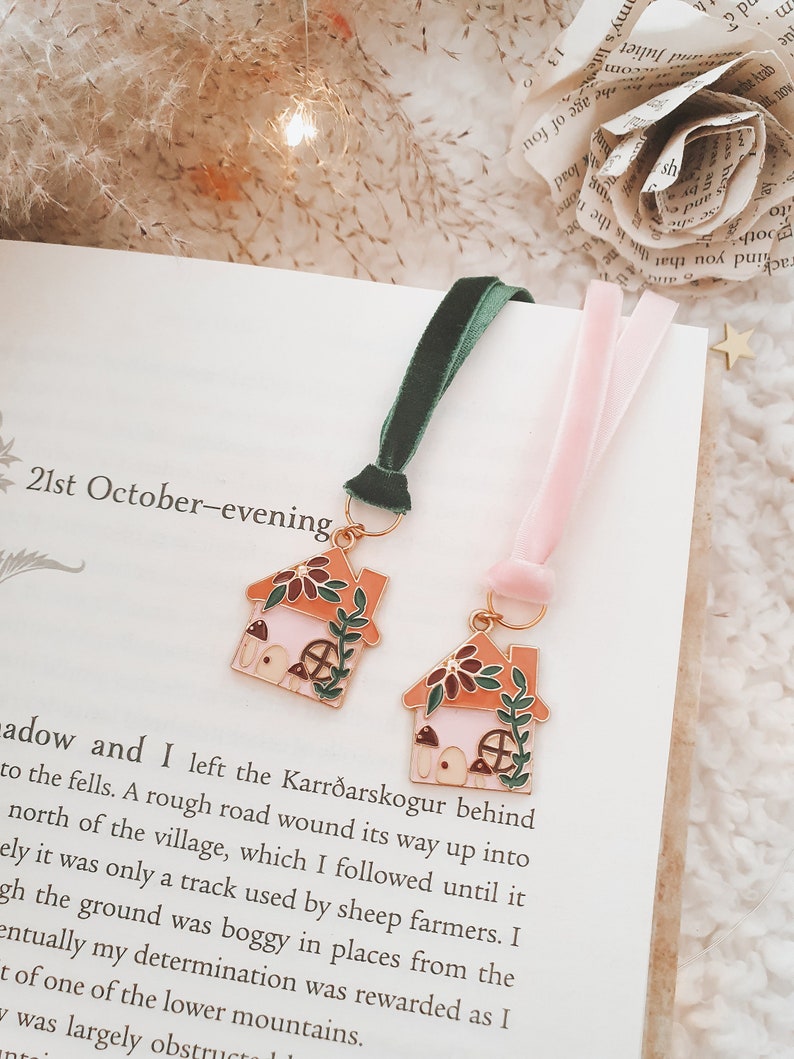 May include: Two gold-toned metal bookmarks shaped like houses with pink and green details. The bookmarks are attached to pink and green velvet ribbons. The bookmarks are on a book with the text "21st October-evening" visible.