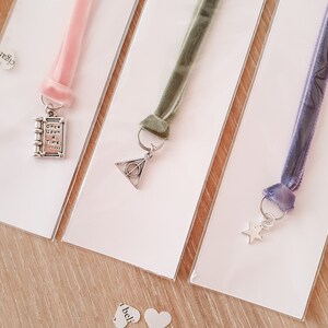 May include: Three velvet ribbon bookmarks with different charms. The first bookmark is pink with a silver charm of a book with the text "Once Upon A Time". The second bookmark is green with a silver charm of the Deathly Hallows symbol. The third bookmark is purple with a silver star charm.