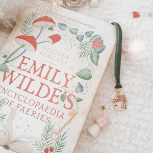 May include: A white book with a green bookmark and a small house charm. The book cover features an illustration of a key, a book, and a red berry with green leaves. The title of the book is "Emily Wilde's Encyclopedia of Faeries".