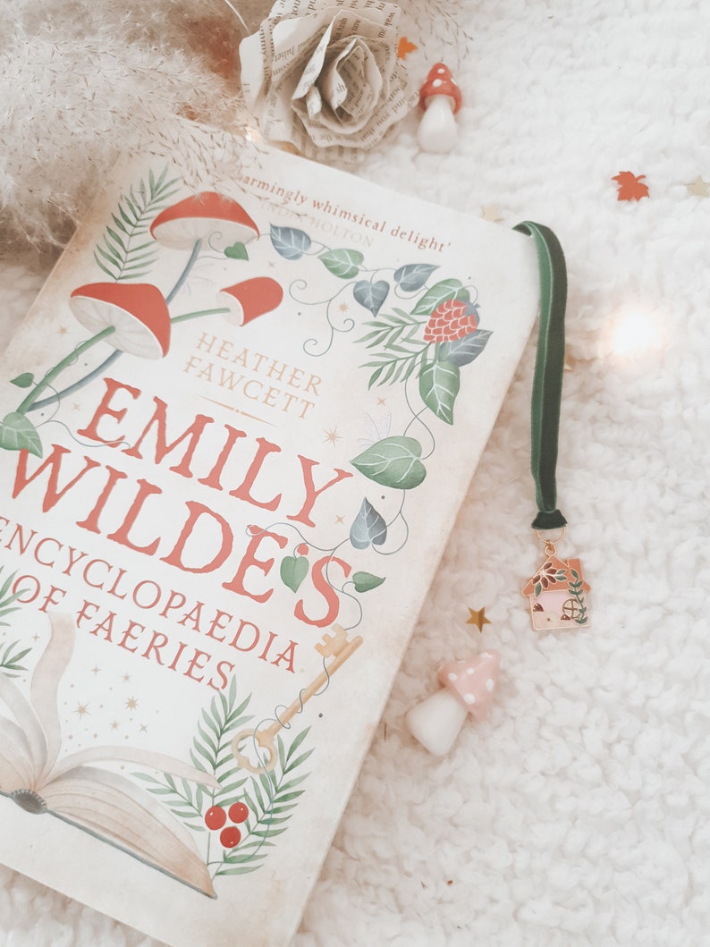 May include: A white book with a green bookmark and a small house charm. The book cover features an illustration of a key, a book, and a red berry with green leaves. The title of the book is "Emily Wilde's Encyclopedia of Faeries".