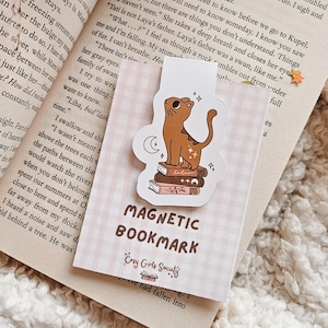 May include: A magnetic bookmark featuring a cartoon cat sitting on a stack of books. The bookmark is pink and white with the text "Magnetic Bookmark" and "Cozy Girls Society" in black.
