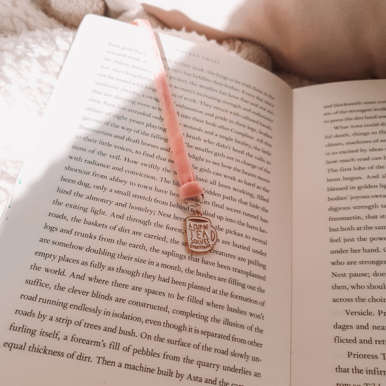 May include: A pink velvet bookmark with a gold charm that reads "A Cup of Tea Solves Everything". The bookmark is resting on an open book with text about a forest.