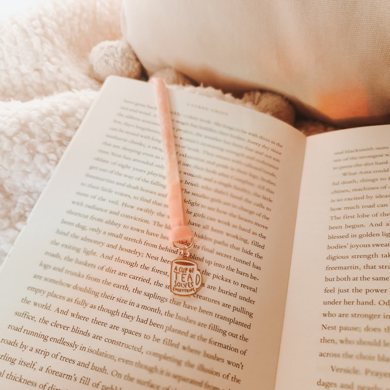 May include: A pink ribbon bookmark with a gold charm that says "A Cup of Tea Solves Everything". The bookmark is placed in an open book with text on the pages.