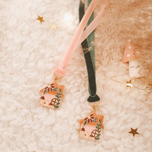 May include: Two pink and orange enamel house charms with green vines and mushroom details, attached to pink and green velvet ribbons.