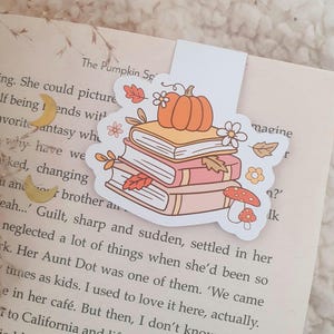 May include: A decorative bookmark with a stack of books, a pumpkin, and autumn leaves. The bookmark is white with a colorful illustration. The design includes orange, pink, and brown elements, ideal for book lovers.