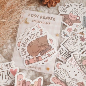 May include: A sticker pack with a pink and white colour scheme featuring illustrations of books, cats, and reading-themed quotes. The sticker pack is labelled "Cosy Reader Sticker Pack" and includes the quote "Read. Dream. Repeat."