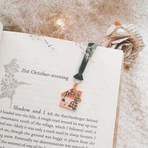 May include: A close-up of a book with a bookmark featuring a small, detailed enamel house charm. The book is open to a page with the text "21st October-evening" and a passage about a journey into the fells.