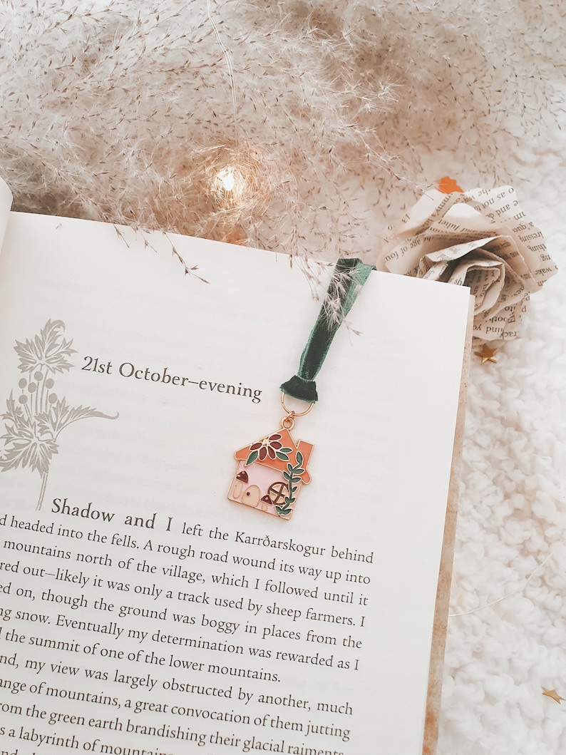 May include: A close-up of a book with a bookmark featuring a small, detailed enamel house charm. The book is open to a page with the text "21st October-evening" and a passage about a journey into the fells.