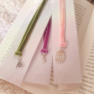 May include: Three velvet ribbon bookmarks with different charms. The first bookmark is green with a silver Harry Potter symbol charm. The second bookmark is purple with a silver star charm. The third bookmark is pink with a gold charm of a teacup that says "A Cup of Tea Solves Everything."