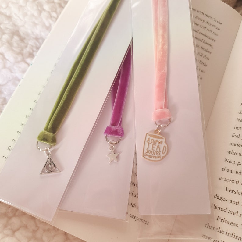 May include: Three velvet ribbon bookmarks with different charms. The first bookmark is green with a silver Harry Potter symbol charm. The second bookmark is purple with a silver star charm. The third bookmark is pink with a gold charm of a teacup that says "A Cup of Tea Solves Everything."