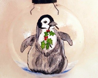 May include: A hand-painted glass ornament featuring a penguin holding a sprig of holly with red berries. The ornament is clear glass with a black top and a white ribbon.