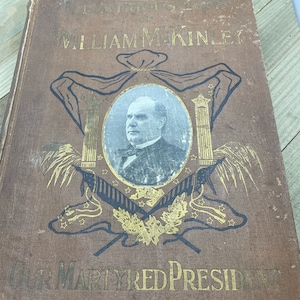 Puede incluir: Un libro marrón con letras doradas en la portada. El título es "The Life, Trials and Assassination of William McKinley, Our Martyred President". La portada presenta un retrato de William McKinley en un círculo rodeado de estrellas y una cinta.