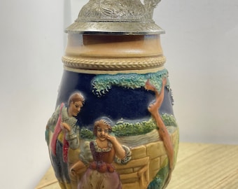 May include: A ceramic beer stein featuring a blue and white scene of a couple in a landscape. The stein has a metal lid with a handle.