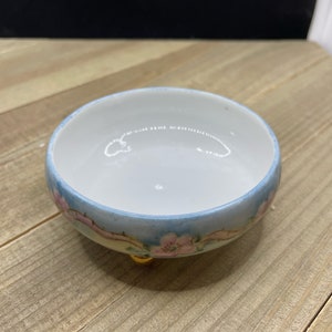 Puede incluir: Un pequeño cuenco de cerámica blanca con un diseño floral azul y rosa. El cuenco tiene tres patas doradas y está sobre una superficie de madera.
