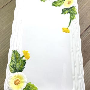 May include: A rectangular white ceramic serving tray featuring a raised, embossed basket-weave border. The tray is decorated with hand-painted yellow flowers and green leaves positioned at the top right and bottom left corners of the surface.