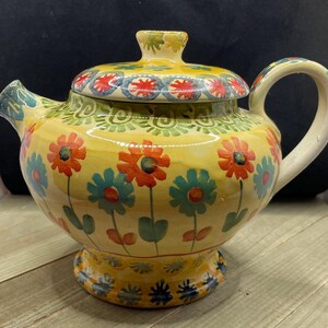 May include: A yellow ceramic teapot with a floral design. The teapot has a lid with a red and blue pattern. The teapot is decorated with red, orange, blue, and green flowers.