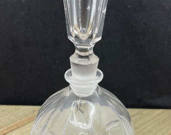 May include: A clear, faceted crystal perfume bottle with a teardrop-shaped stopper. The bottle has a round, ribbed body and a frosted glass stopper. A gold label with black text is affixed to the bottle.