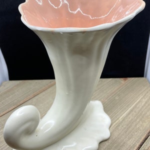 May include: A white ceramic vase shaped like a horn with a pink interior. The vase has a scalloped rim and a curled base.