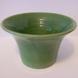 May include: A wide-mouthed ceramic bowl with a glossy, solid green glaze. The vessel features a flared rim that tapers down to a narrow, flat base. The interior and exterior surfaces show subtle, concentric circular ridges from the pottery process.
