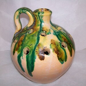 May include: A round ceramic flower frog vase with a single handle and a narrow neck. The beige base features a glossy glaze with vertical drips of vibrant green and dark brown running down from the top, interspersed with multiple circular holes.