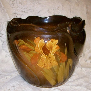May include: A dark brown ceramic planter with a glossy finish and a wavy, scalloped rim. The front features a hand-painted, raised relief design of a large, golden-yellow iris flower with green leaves, set against the deep brown background.