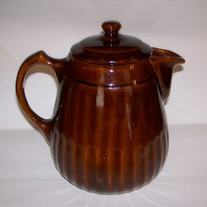May include: A glossy dark brown ceramic teapot featuring a vertical ribbed texture along the body, a matching lid with a small knob handle, a curved side handle, and a pointed pouring spout. The teapot has a smooth, reflective finish.