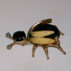 May include: A vintage insect brooch with a gold-tone metal body. The brooch features a black, oval-shaped body with gold-colored wings and legs. The head has a black stone and the legs have small blue stones.
