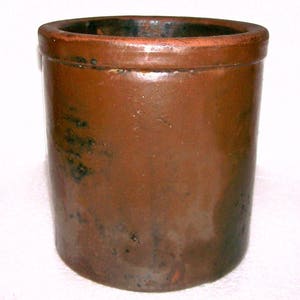May include: A cylindrical brown stoneware crock featuring a smooth, glossy glaze with subtle dark mottled spots and a thick, rounded rim. The vessel has a deep, open interior and a flat base, showcasing a uniform, sturdy construction throughout.
