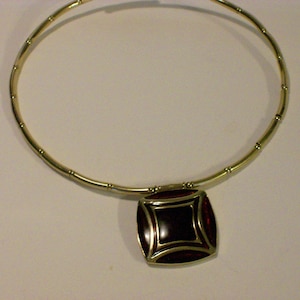 May include: A gold-toned necklace with a square pendant. The pendant features a dark red center framed by a gold border and a red outer edge. The necklace has a rigid, circular design.