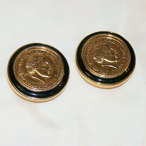May include: Two gold-toned coin-shaped earrings with black enamel borders. The coins feature a profile of a person and text around the edge. The earrings have a gold-colored frame.