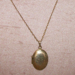 May include: A gold-colored oval locket pendant on a delicate gold chain. The locket has a simple, classic design and hangs from a slender chain. The background is a soft, neutral color.