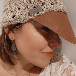 Peut inclure: Une femme portant un chapeau crocheté beige avec une visière en cuir marron et des boucles d'oreilles argentées. Elle porte un haut en dentelle blanche et un collier en or avec des pendentifs triangulaires.