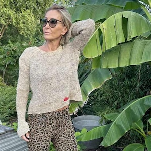 Könnte beinhalten: Ein beiger Strickpullover mit einem roten Lippen-Design. Der Pullover hat eine lockere Passform und eine leicht verkürzte Länge. Das Model trägt den Pullover zu einer Hose mit Leopardenmuster.