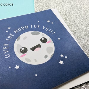 May include: Greeting card with a navy blue background featuring a smiling cartoon moon with pink cheeks and the text "OVER THE MOON FOR YOU!". White stars are scattered around the moon. The card is on a white surface.