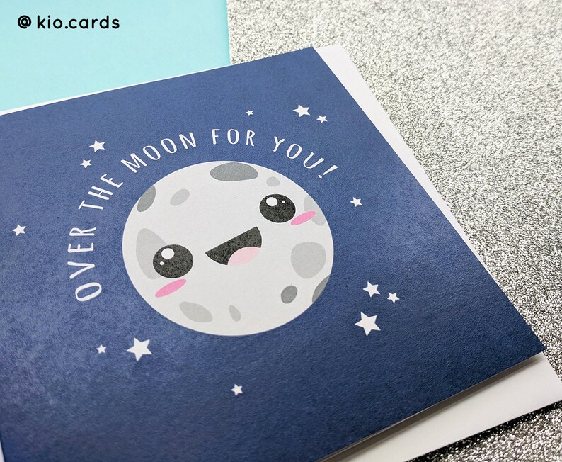 May include: Greeting card with a navy blue background featuring a smiling cartoon moon with pink cheeks and the text "OVER THE MOON FOR YOU!". White stars are scattered around the moon. The card is on a white surface.