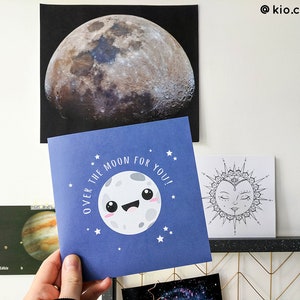 May include: A blue greeting card with a smiling moon illustration and the text "OVER THE MOON FOR YOU!". The card is held in front of a large moon print and other space-themed art. The overall theme is space and celestial bodies.