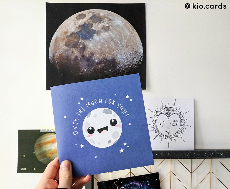 May include: A blue greeting card with a smiling moon illustration and the text "OVER THE MOON FOR YOU!". The card is held in front of a large moon print and other space-themed art. The overall theme is space and celestial bodies.