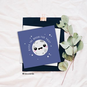 May include: A square greeting card with a smiling moon illustration and the text "OVER THE MOON FOR YOU!" on a navy blue background. The card is surrounded by a white envelope, a black card, and sprigs of greenery. The card has a cute, cartoonish design.