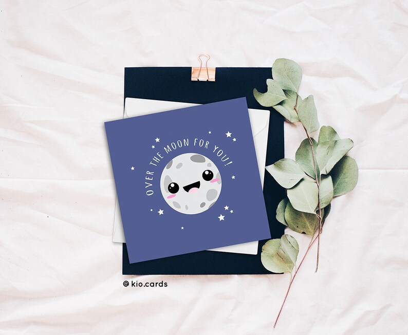 May include: A square greeting card with a smiling moon illustration and the text "OVER THE MOON FOR YOU!" on a navy blue background. The card is surrounded by a white envelope, a black card, and sprigs of greenery. The card has a cute, cartoonish design.