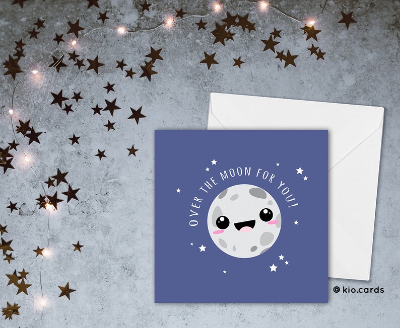 May include: A square greeting card with a cartoon moon illustration on a blue background. The text "OVER THE MOON FOR YOU!" surrounds the moon. A white envelope is next to the card. The background has star-shaped confetti.
