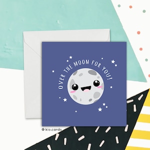 May include: A square greeting card with a cartoon moon illustration on a dark blue background. The text "OVER THE MOON FOR YOU!" is written around the moon. A white envelope is behind the card. The card has a cute, whimsical design.