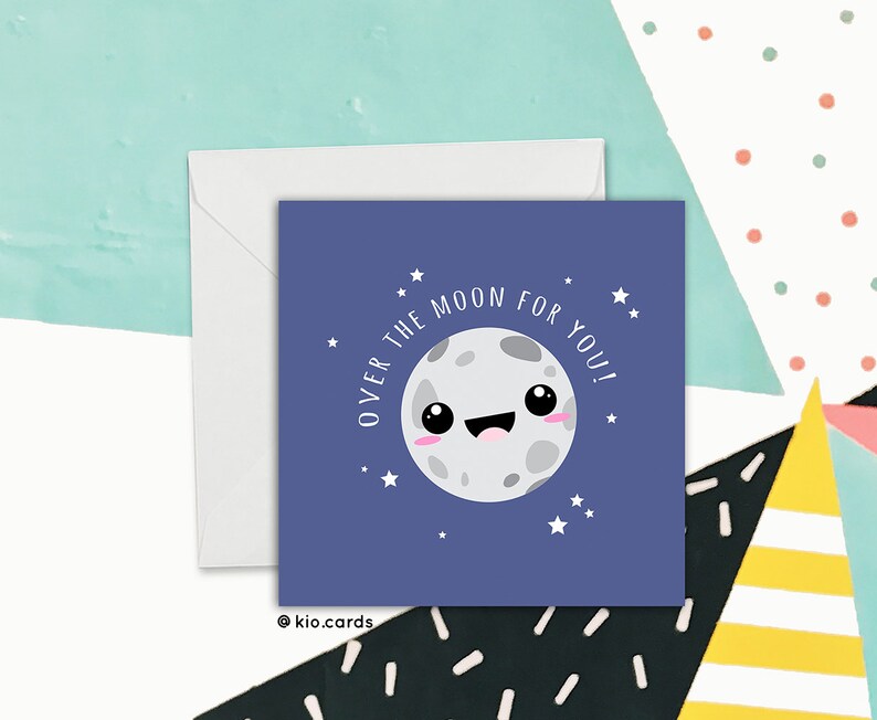 May include: A square greeting card with a cartoon moon illustration on a dark blue background. The text "OVER THE MOON FOR YOU!" is written around the moon. A white envelope is behind the card. The card has a cute, whimsical design.