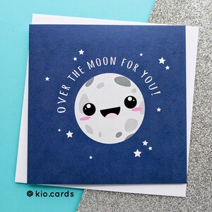 May include: A navy blue greeting card with a smiling cartoon moon illustration and the text "OVER THE MOON FOR YOU!". The card is surrounded by white stars and a white border. The bottom left corner has the text "@kio.cards".