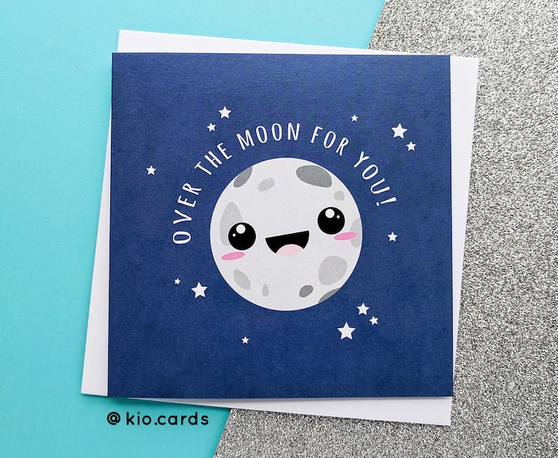May include: A navy blue greeting card with a smiling cartoon moon illustration and the text "OVER THE MOON FOR YOU!". The card is surrounded by white stars and a white border. The bottom left corner has the text "@kio.cards".