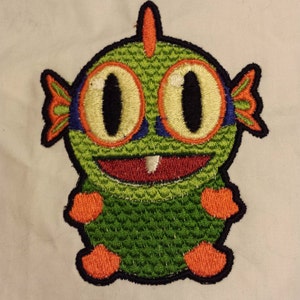 以下が含まれることがあります： 緑、オレンジ、青の刺繍パッチ。大きな目と歯を見せて笑っている漫画のモンスターです。
