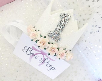 Puede incluir: Corona de fieltro blanco con un número uno de purpurina plateada y una guirnalda de flores rosas. La corona tiene una cinta de satén blanco. El texto "Bow Peep" está escrito en una tarjeta blanca.