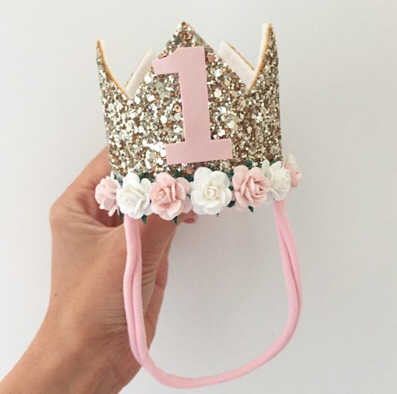 May include: Gold glitter crown with a pink felt number one and a pink headband decorated with white and pink flowers.