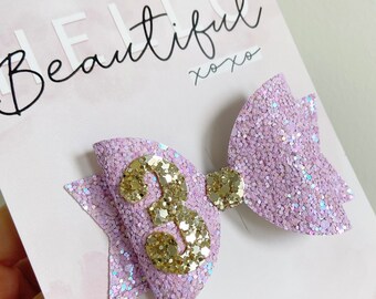May include: A light purple glitter bow with a gold glitter number 3 in the centre. The bow is on a card that says "Hello Beautiful xoxo".